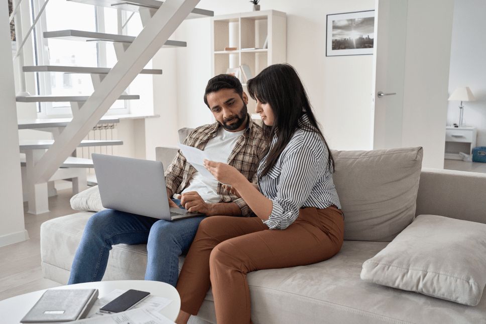 A man and woman on a couch looking at a paper and laptop.