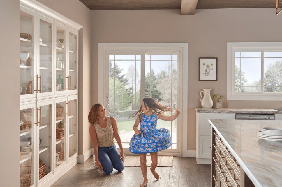Woman smiling at child dancing in the interior kitchen of a home in front of white sliding patio doo